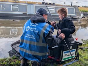A lets fish coach alongside a young boy teaching him how toi fish on the canalside with a canalboat in the background