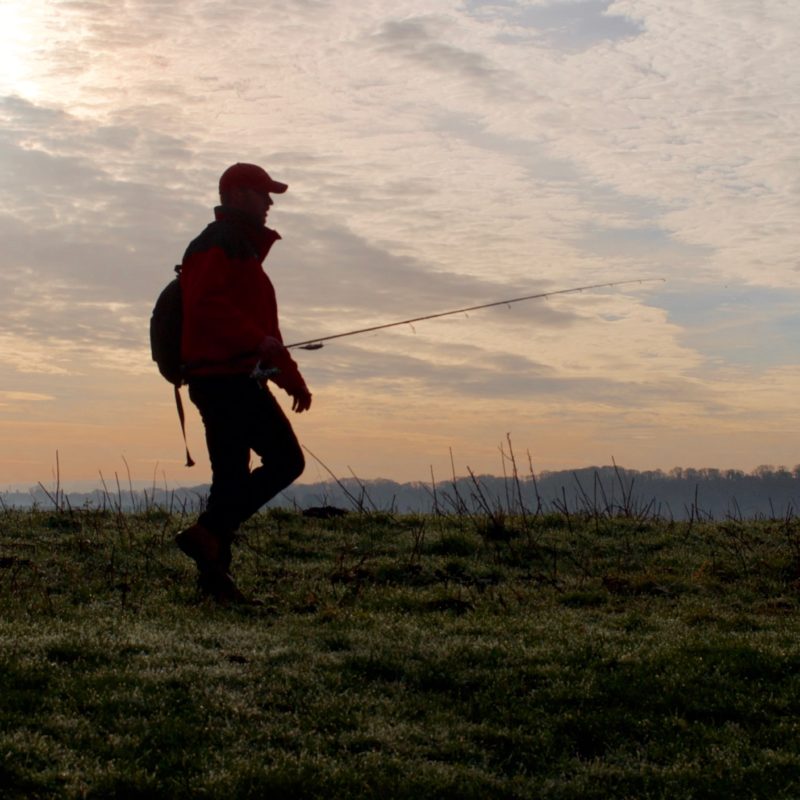 a solo angler walking along the riverbank with rod in hand on a crisp autumn morning