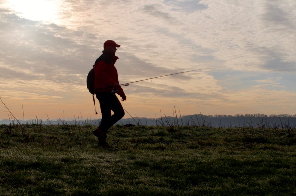 a solo angler walking along the riverbank with rod in hand on a crisp autumn morning